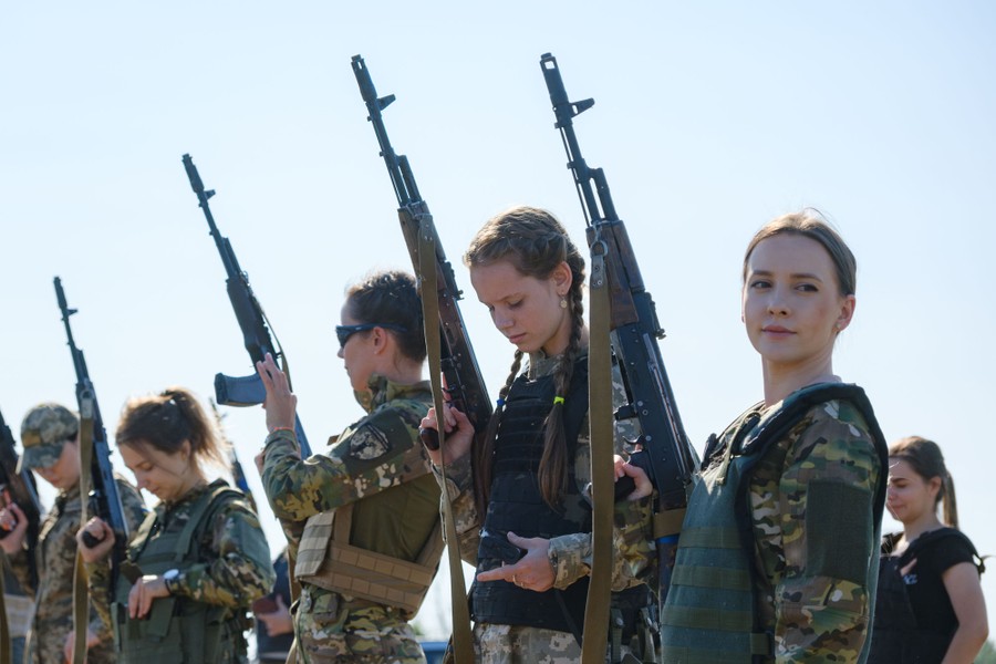 A half dozen women wearing varying pieces of combat gear stand side-by-side, holding weapons.