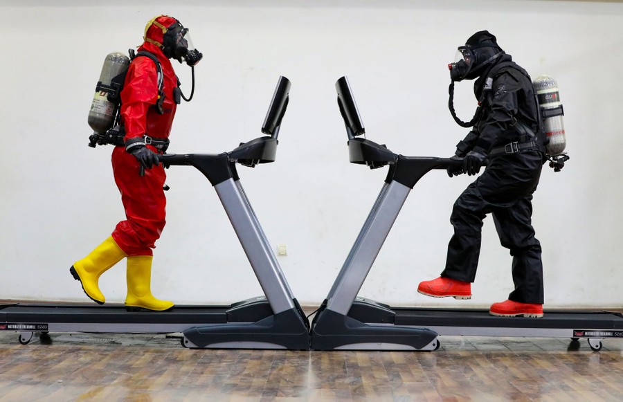 Two people wearing heavy protective gear and oxygen masks face each other while walking on treadmills.