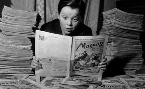 A black-and-white photo of a boy wearing a black shirt and peering at a newspaper comic, with newspapers stacked on both sides