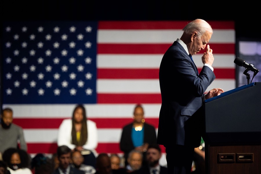 President Joe Biden leans his head slightly in prayer, standing at a podium, seen from the side, with an audience in the background.