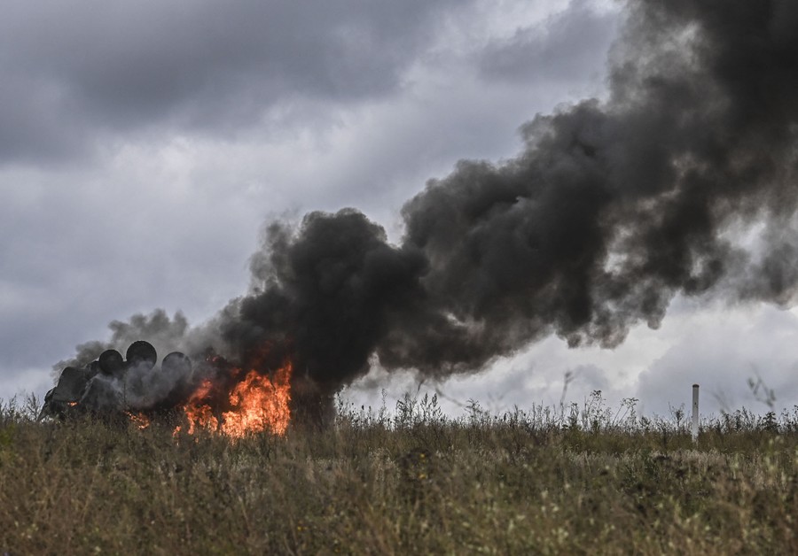 A destroyed military vehicle burns in a field.