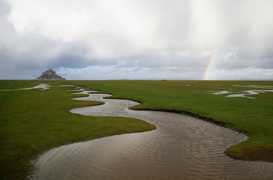 A broad view of a meandering channel in a grassy marsh, with a clutch of buildings on a small island in the distance.