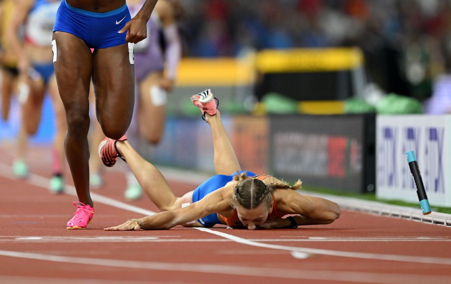 A runner falls to the ground during a relay race, passed by another racer.