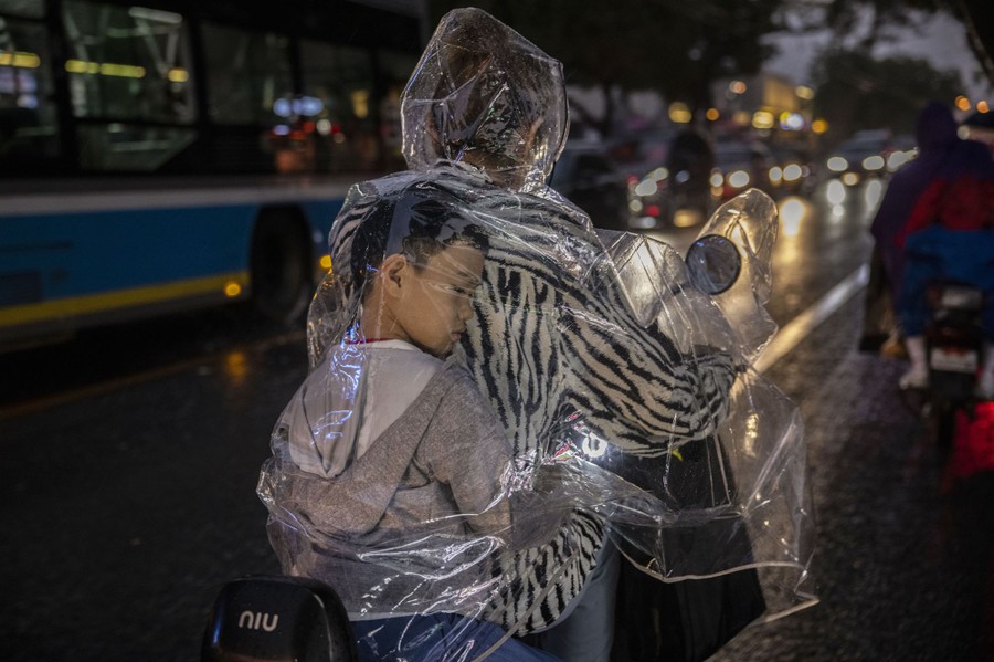 A boy sits behind his mother, inside her clear plastic raincoat, as they ride a scooter.