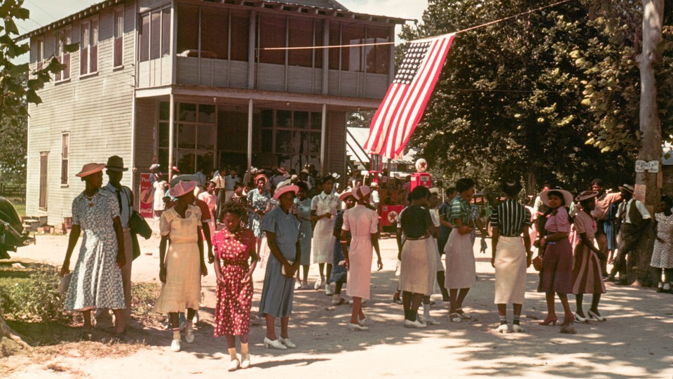 A photograph of group of Black people dressed in colorful summer celebration clothes in front of a hanging American flag and a two-story building
