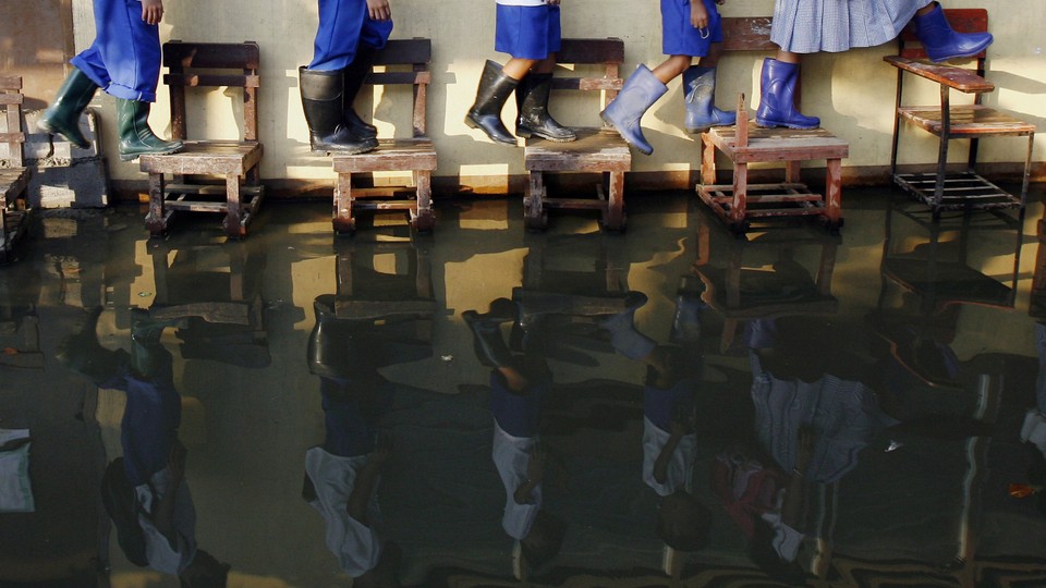 Students wearing rain boots walk on desks across their flooded classroom.