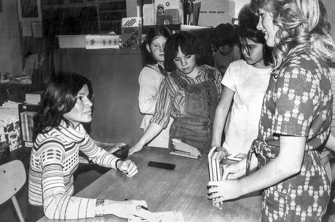 black-and-white photo of Blume in turtleneck with long dark hair seated at table during book signing surrounded by standing girls