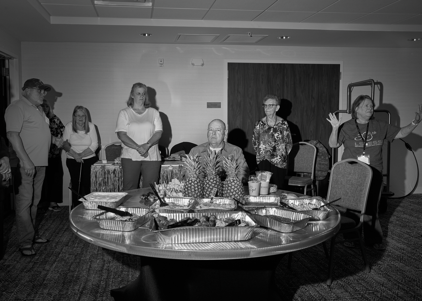 black-and-white photo of group standing around man seated at round table that has several pineapples and trays