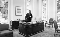 Henry Kissinger standing at a desk in an office with high windows in 1970