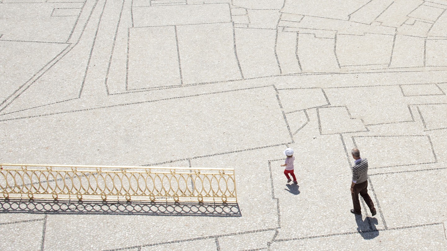 A man walks behind a child who is running across a white landscape with faint black lines in various gridded shapes