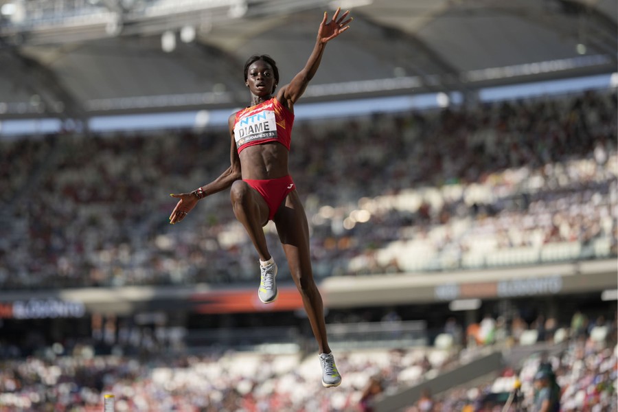 An athlete leaps during a long-jump attempt, with crowded stadium seats in the background.