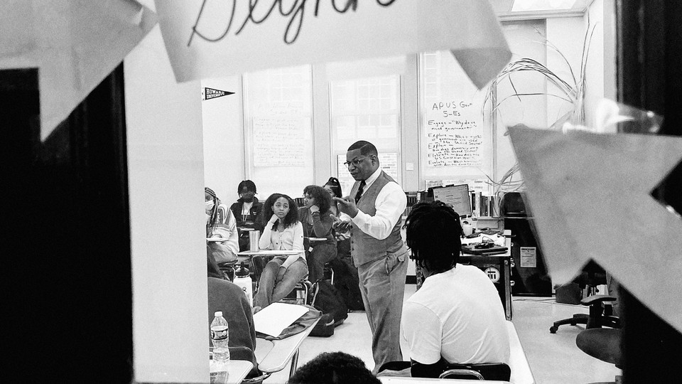 A black-and-white photo of a teacher teaching a class, captured through the window in the classroom door