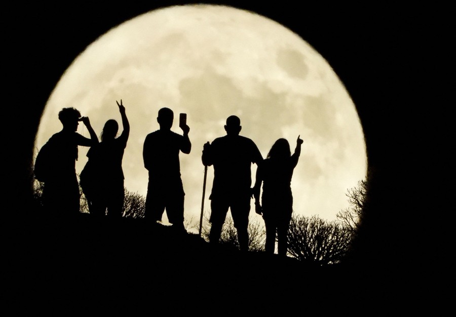 Five people stand on a hillside silhouetted by the full moon.