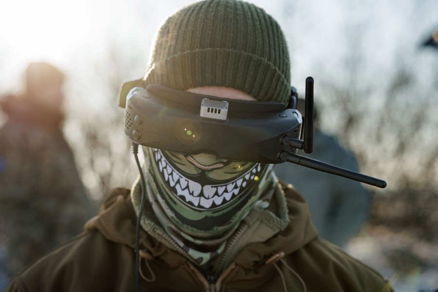 A soldier wears a face covering, a beanie, and a black video headset with antennas poking out of it
