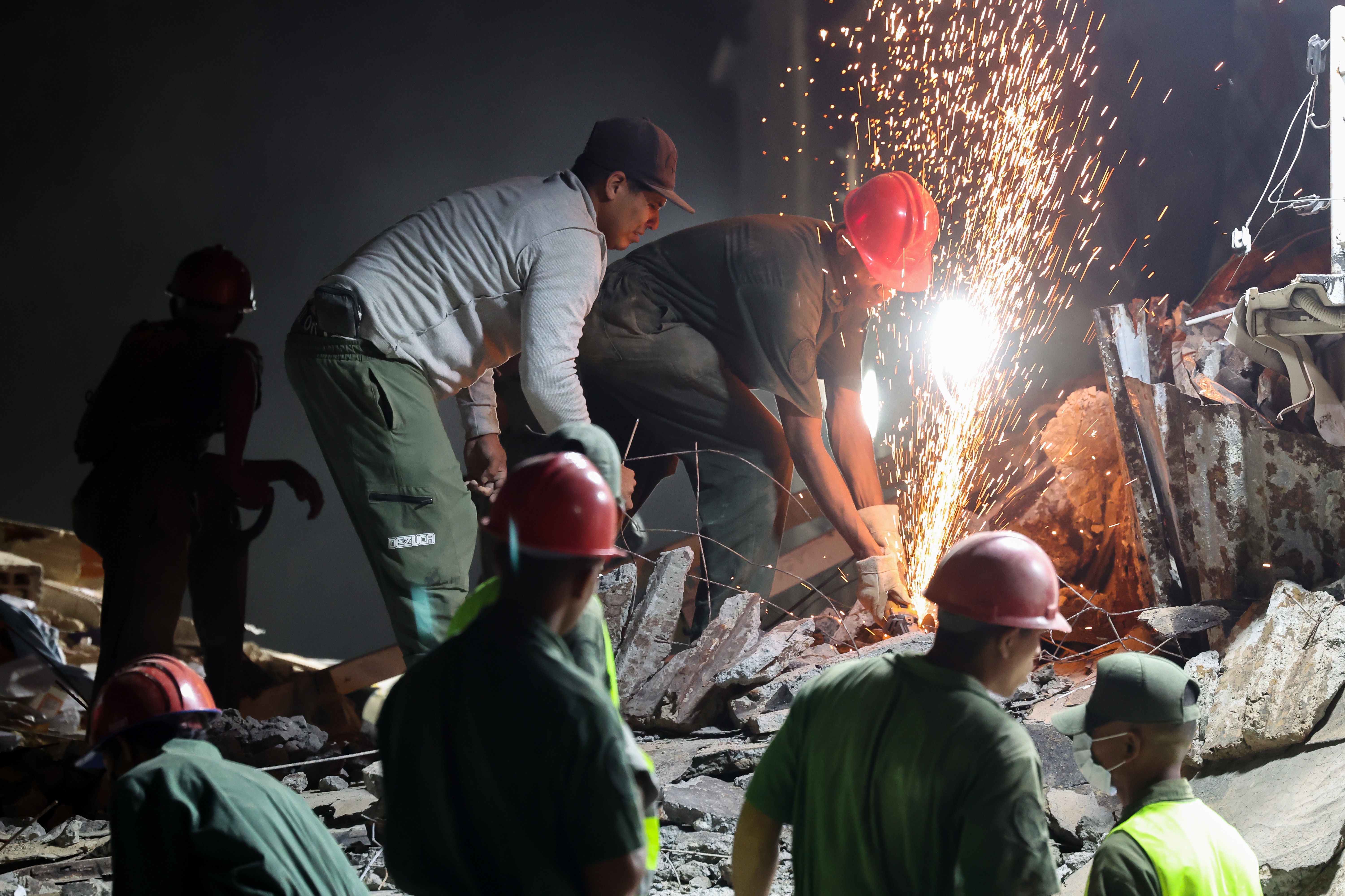 Sparks fly as a rescue worker uses a cutter to break apart metal reinforcing bars in a pile of rubble.