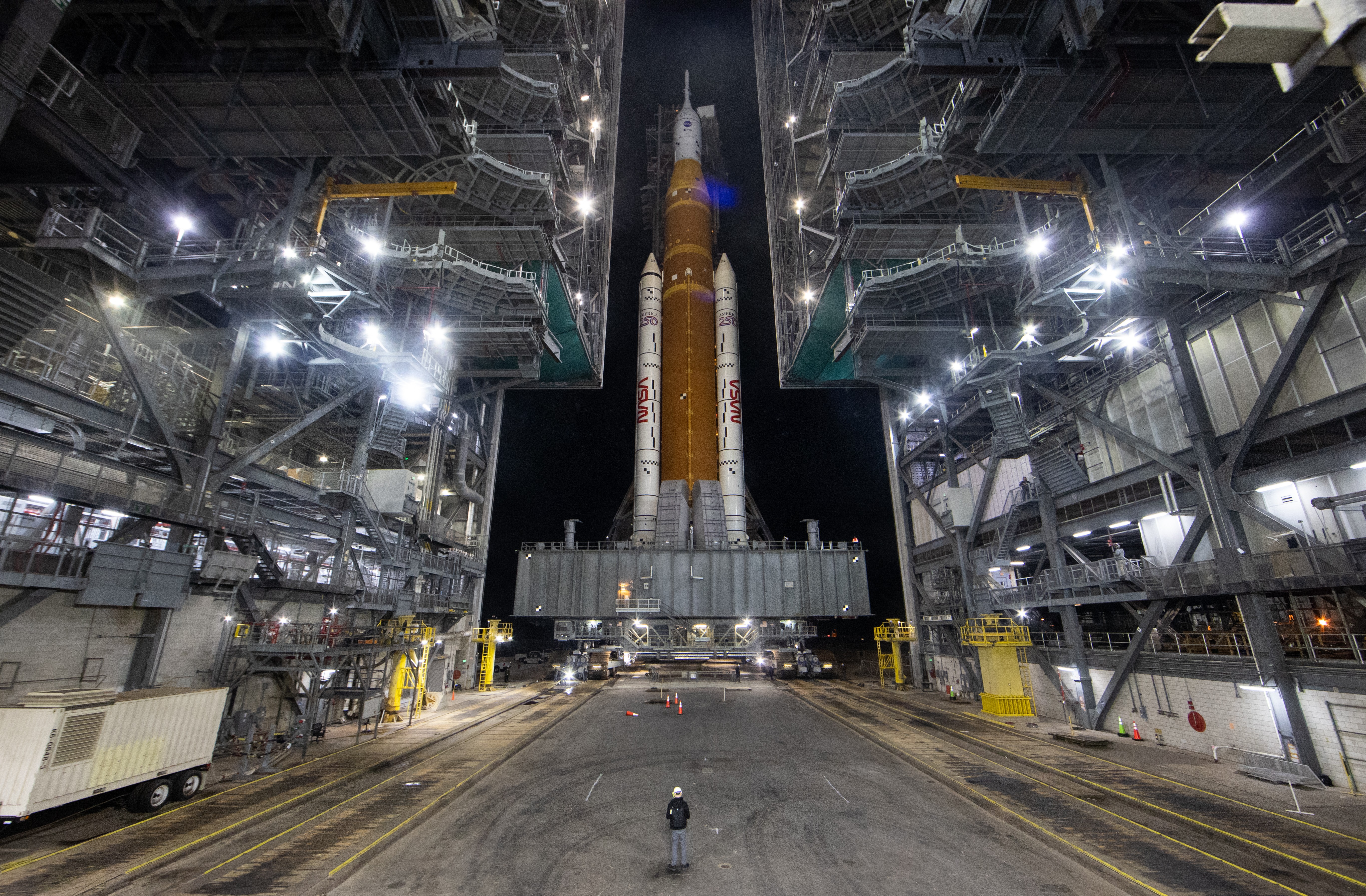 A tall rocket is rolled out of a launch facility building on top of a large tracked vehicle.