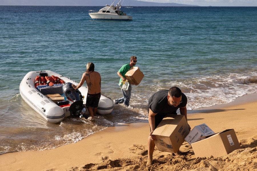 Several people unload boxes onto a beach from a small boat.