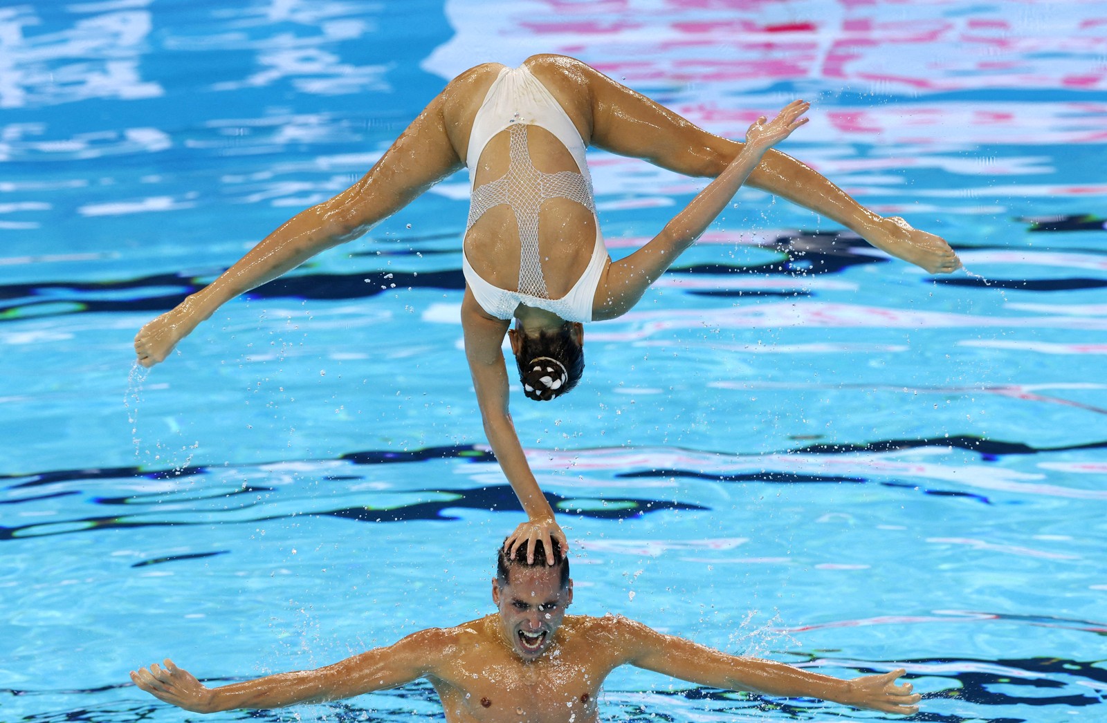 A pair of artistic swimmers seen during a performance, one of them, balancing on one hand placed on the head of the other, who is swimming in a pool.