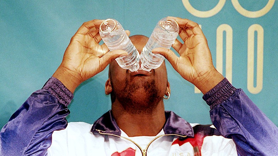 A man drinks from two plastic water bottles simultaneously. Both bottles are tipped over his mouth, blocking the upper half of his face.