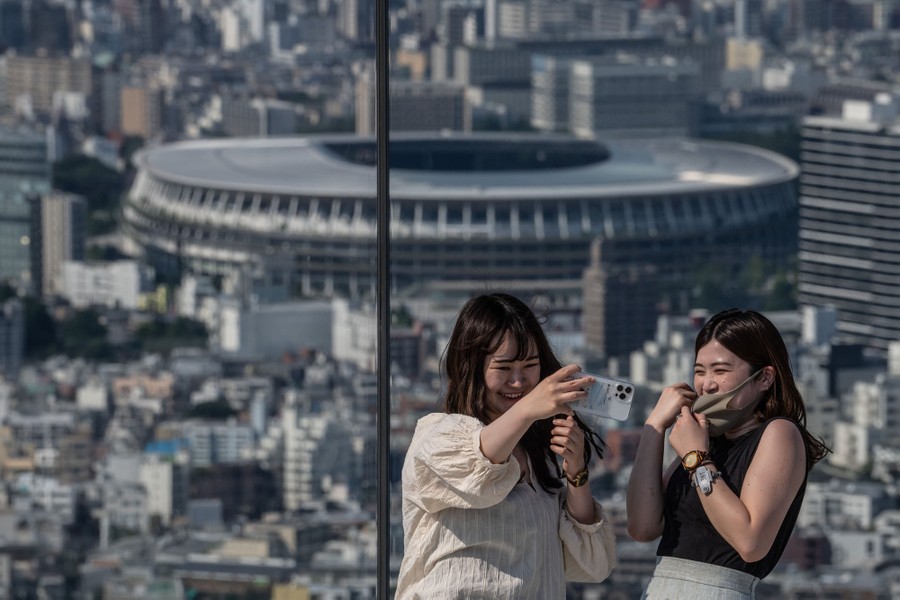 Women take a selfie atop a tall building with the Tokyo Olympic stadium visible in the background.