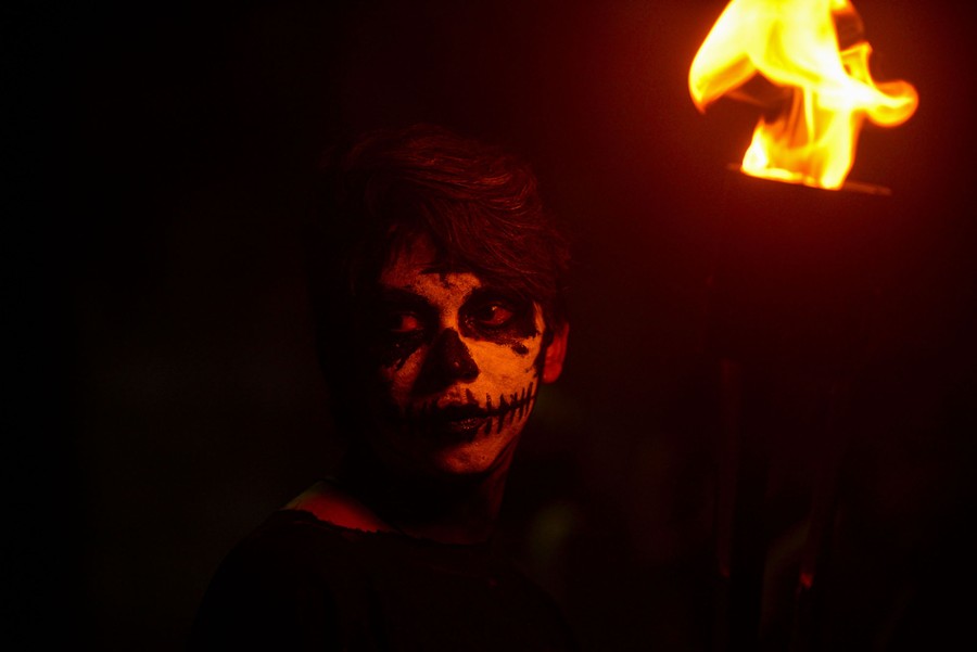 A young person with skull makeup on their face stands near a torch at night.