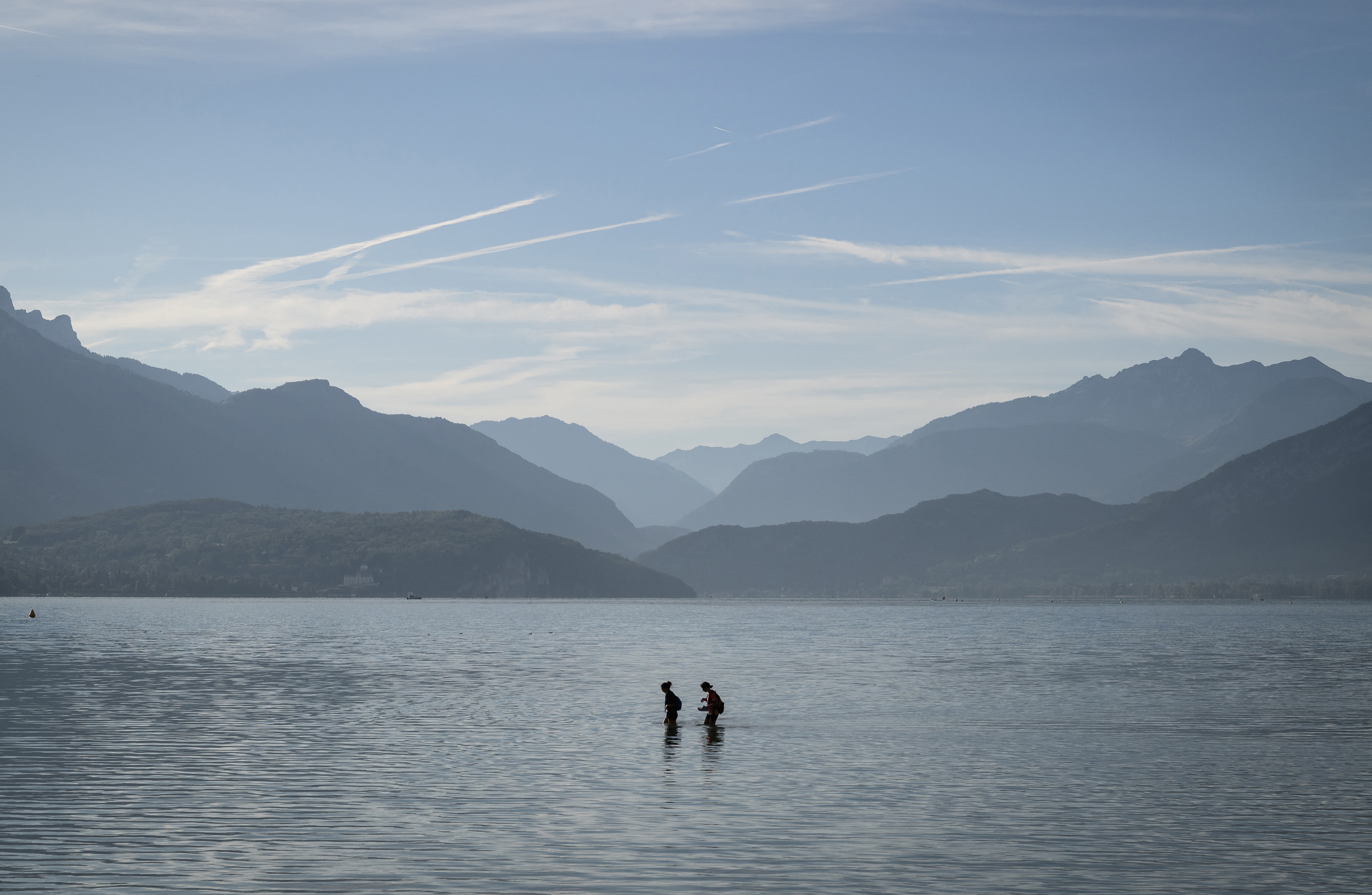 A wide view of two people wading through shallow water in a broad lake, with mountains visible in the background