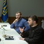 Several people sit around a large table framed by two blue-and-yellow flags; Volodymyr Zelensky speaks while several others take notes.