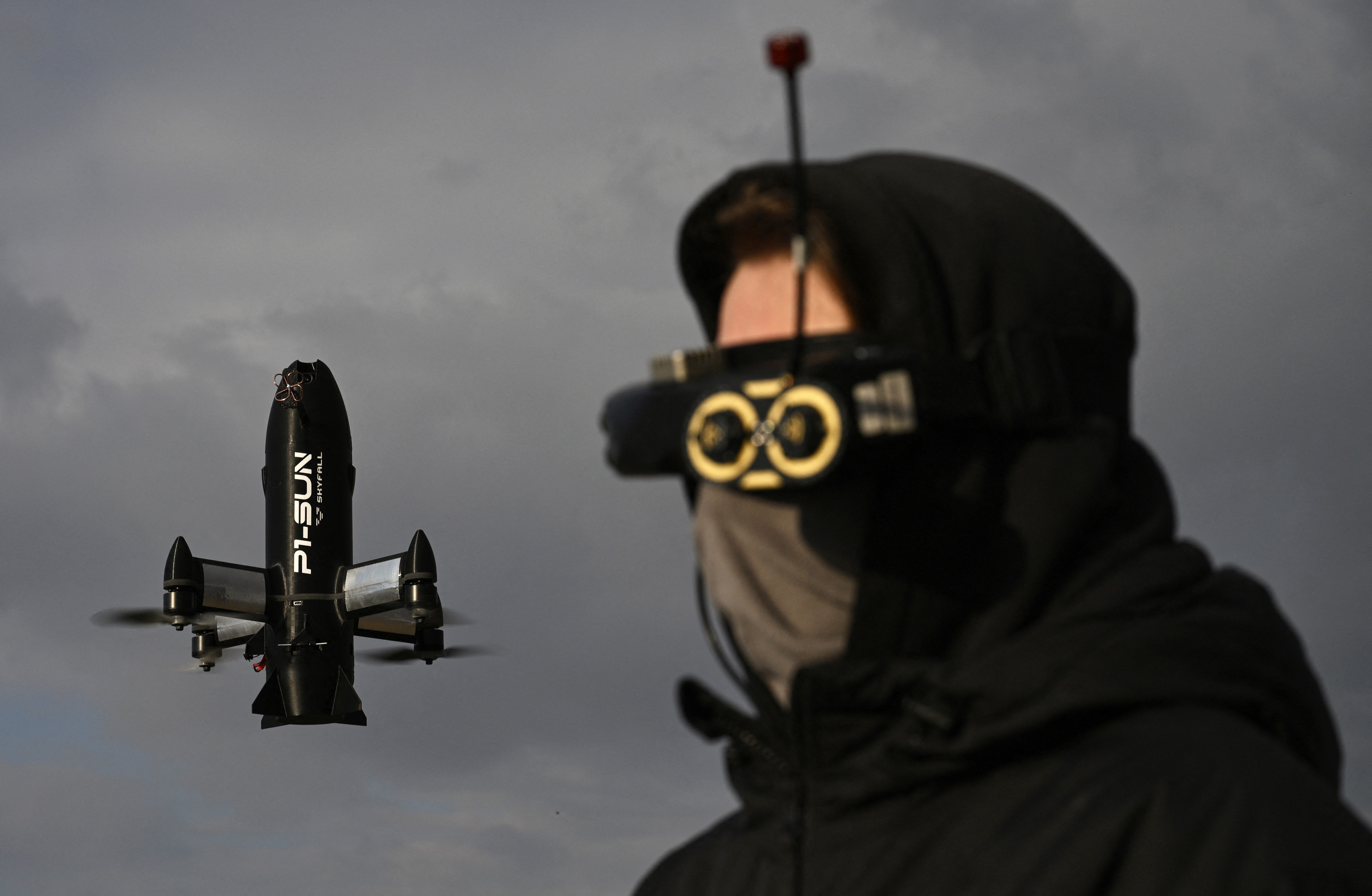 A drone operator, wearing a mask and visor, pilots a drone which hovers in the background.