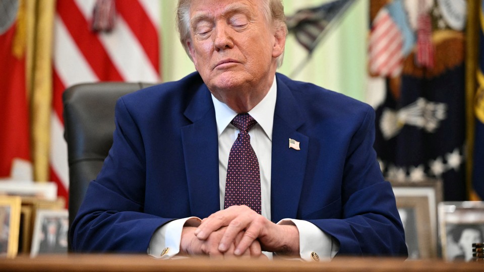 A photo of Trump sitting at the Resolute Desk looking down with clasped hands