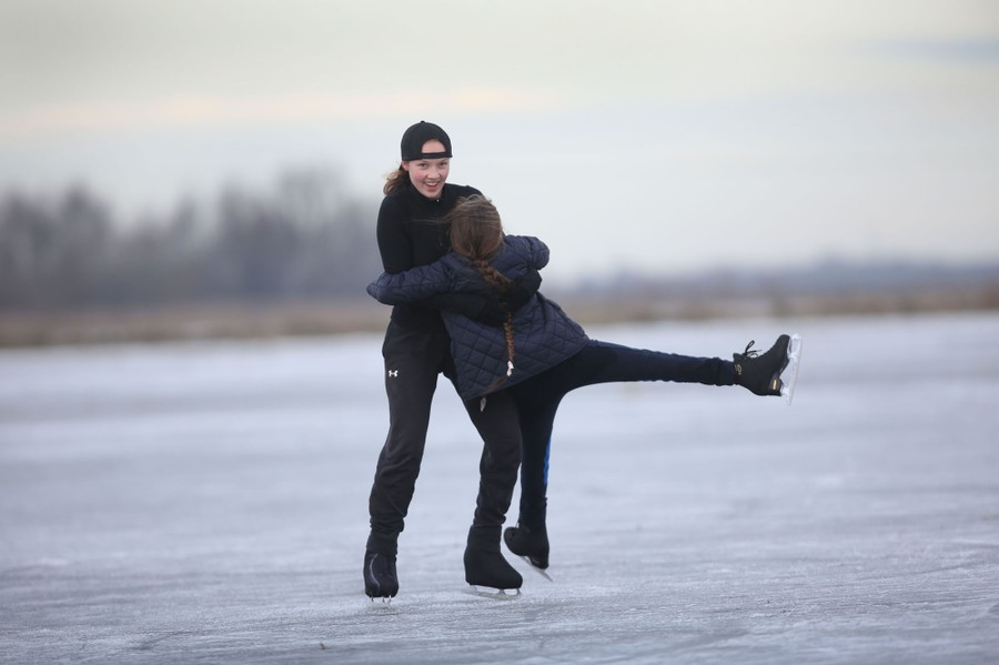 Two girls hold each other, playfully skating on a large patch of ice outside.