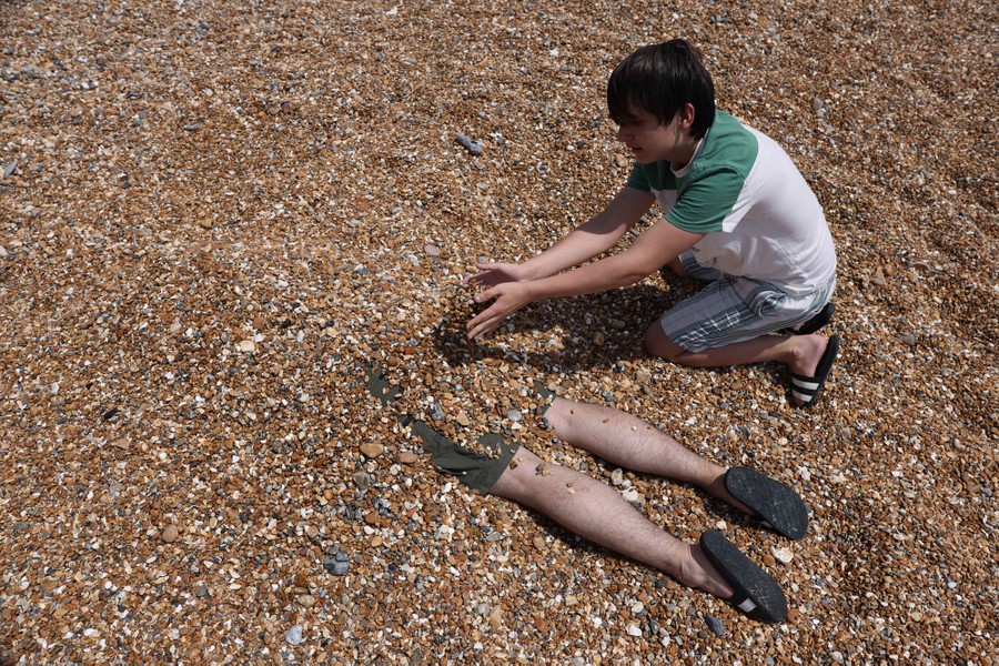 A young person piles small rocks on top of another, mostly buried person on a beach.
