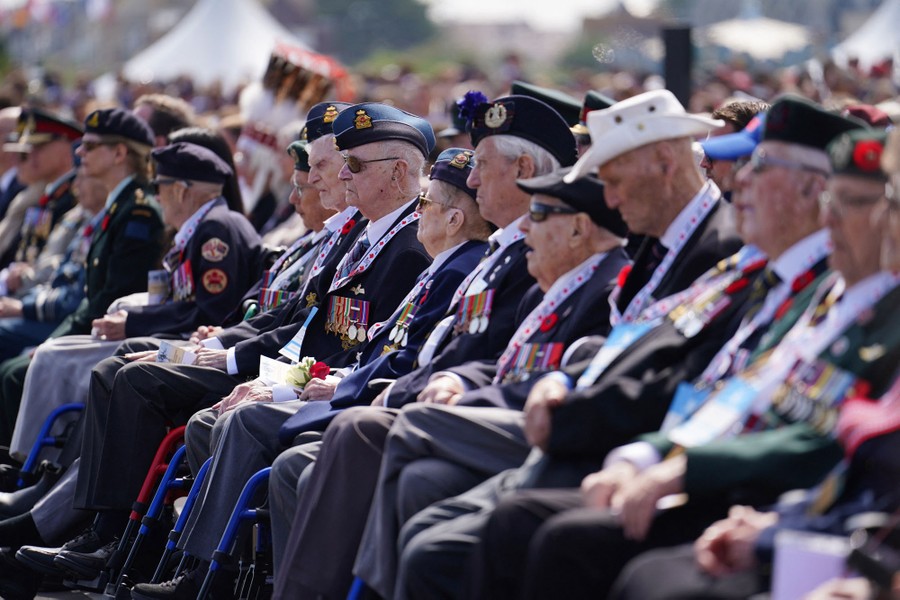A row of older veterans sitting in wheelchairs, in the front row of a large crowd.