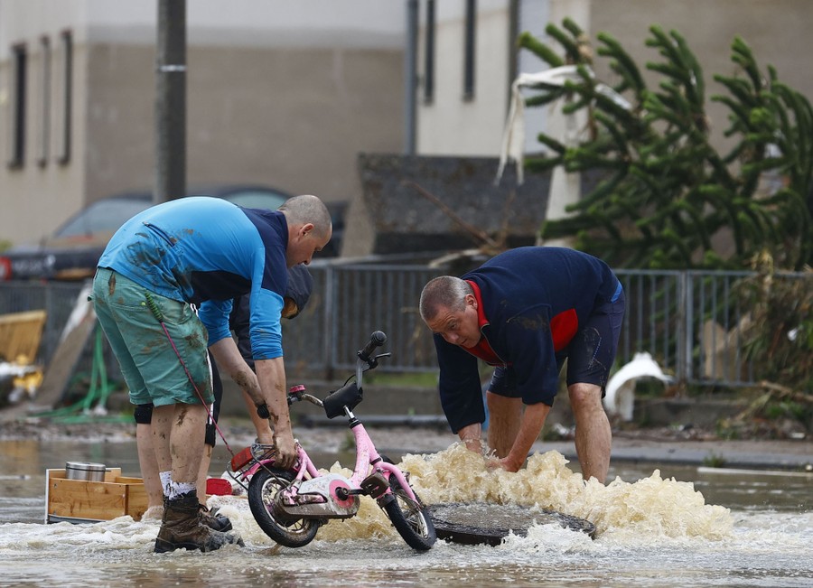 Several people struggle with rising floodwater near a manhole in a street.