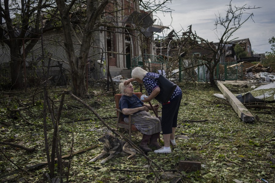 A woman sits in a chair outside of heavily damaged homes, surrounded by leaves and debris. Another woman tends to the first, while a dog rests beside them.