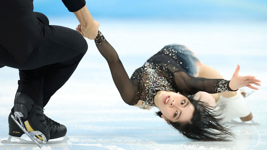 A figure skating pair spins on the ice, with one member low to the ice, being swung around by the other.