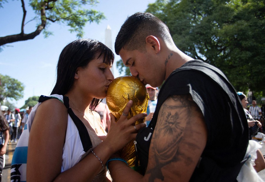 A couple embraces a gold trophy between them, leaning in to kiss it on either side.