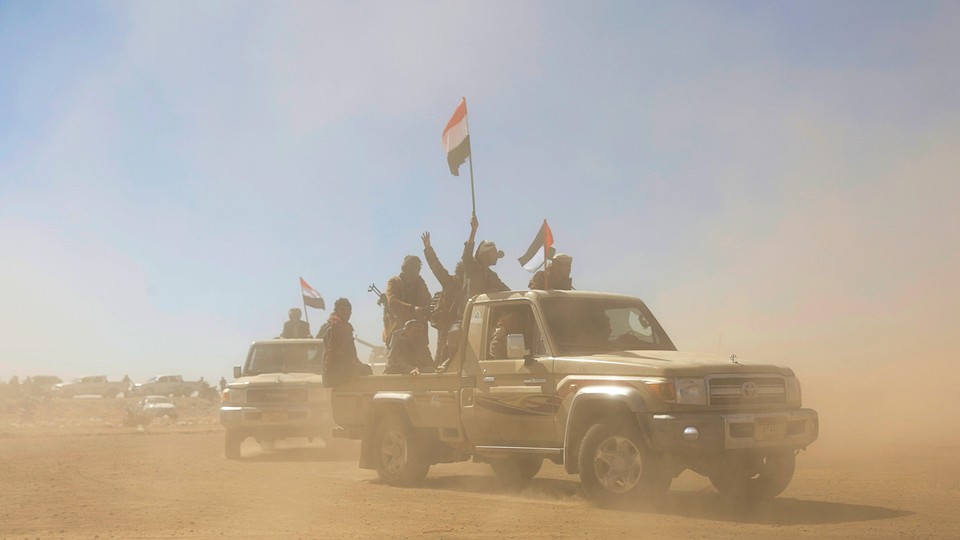 Houthi fighters and tribesmen rally with flags in the back of a pickup truck surrounded by desert dust.