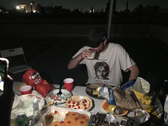 A man sitting on a dark rooftop, drinking a glass of milk, surrounded by party snacks.