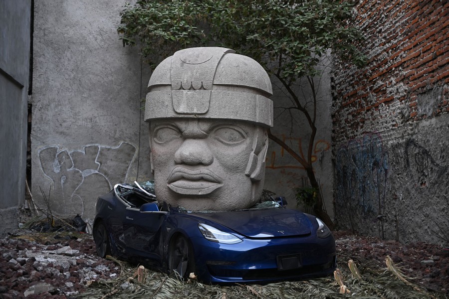 A giant head sculpture sits atop a crushed car in a small lot surrounded by brick and concrete walls.
