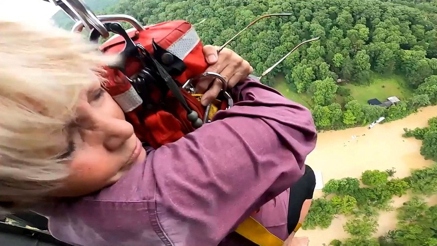 A close-up view of a person being airlifted into a helicopter. Below her lies greenery and a flooded river.