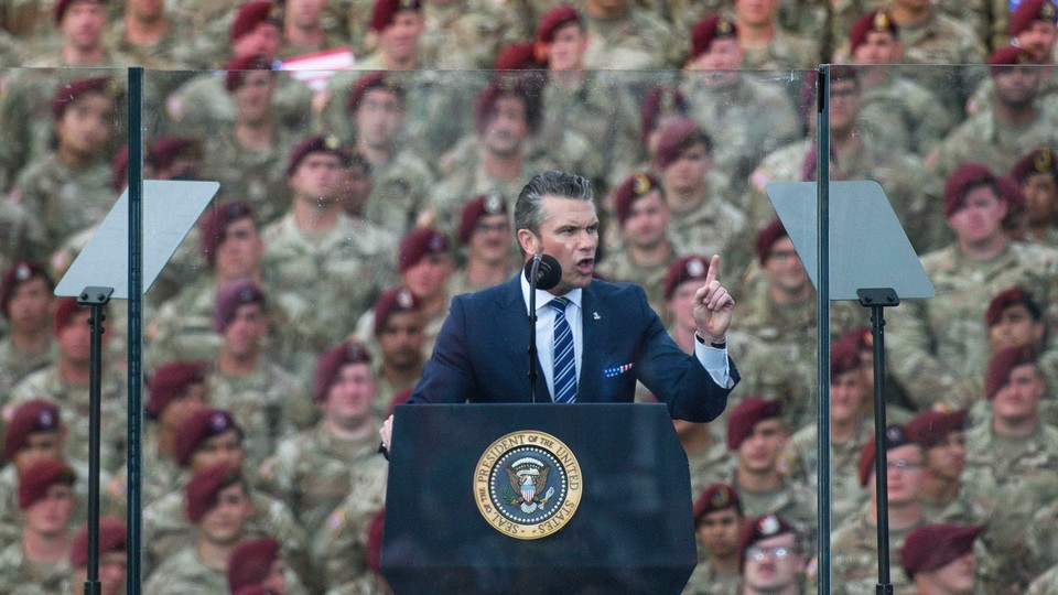 Secretary of Defense Pete Hegseth giving a speech with teleprompters behind a glass barrier, while a crowd of uniformed troops stand in the background