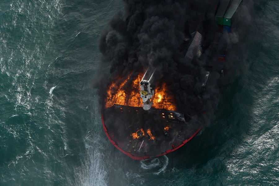 Fire and black smoke rise from a damaged ship, as seen from above.