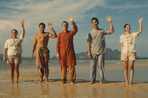 Five people stand waving on a beach