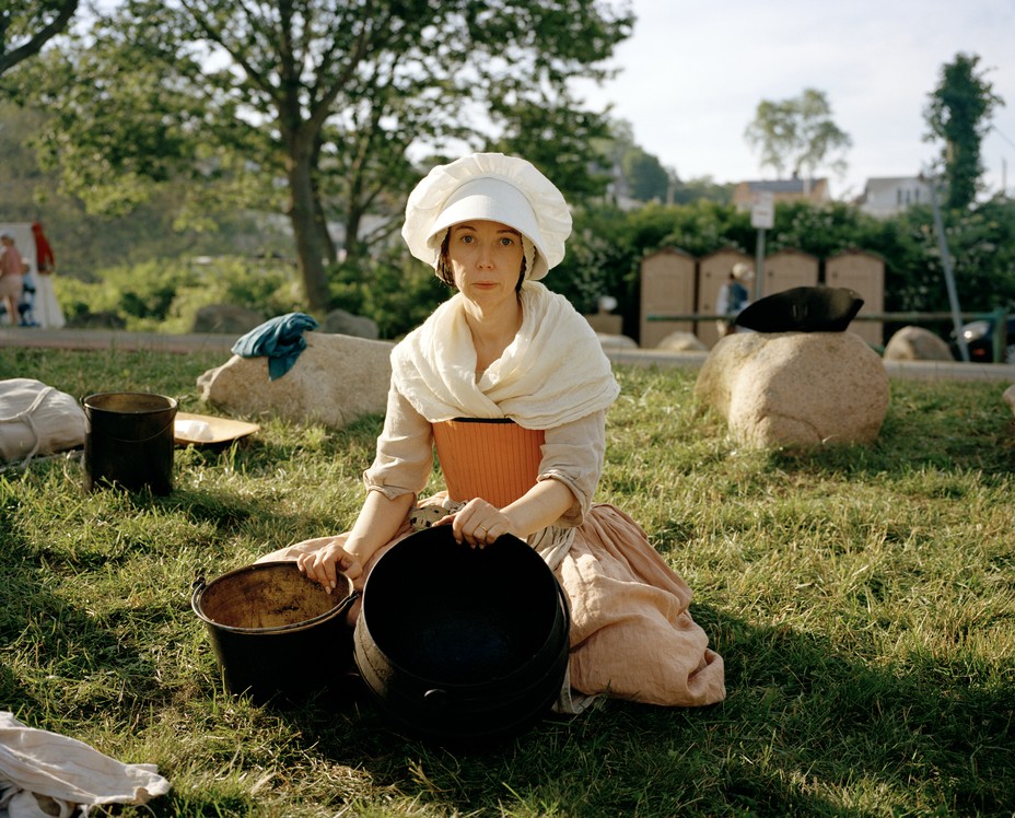 photo of a woman in white colonial bonnet, white shawl, and pink dress kneeling in grass holding two baskets while doing the washing at a reenactment of Bunker Hill