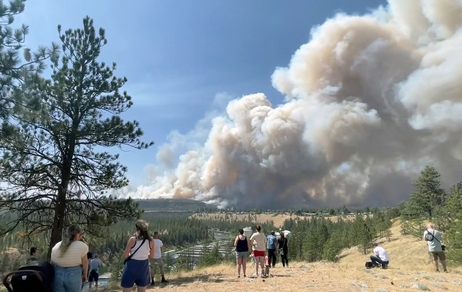 People watch from a bluff above a river as massive plumes of smoke rise from a wildfire.
