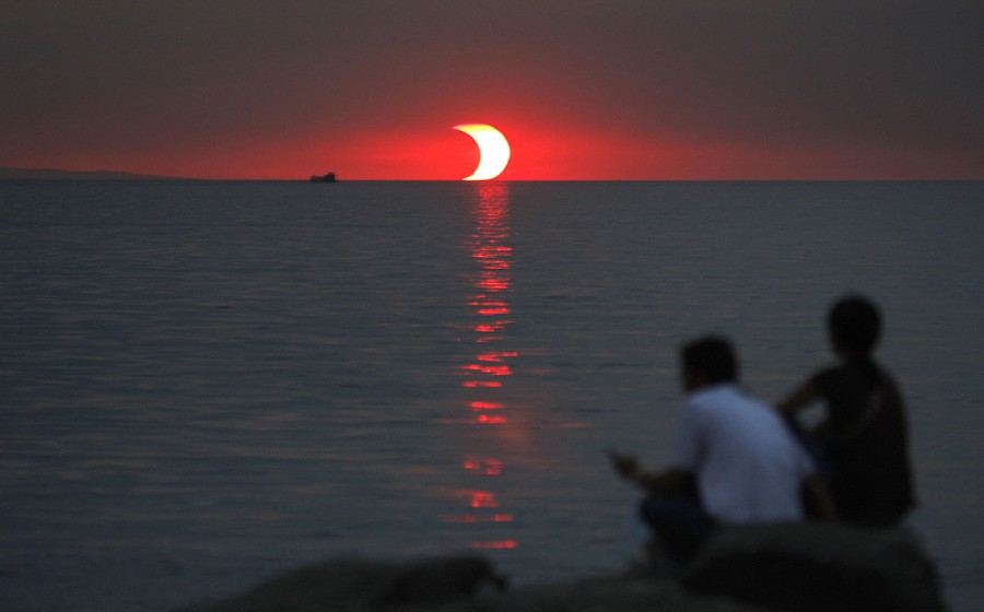 Two people sit on rocks overlooking water, with a partly-eclipsed sun setting in the distance.