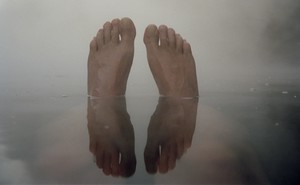 A pair of feet coming out of the water, against a foggy grey sky, and reflected in the water