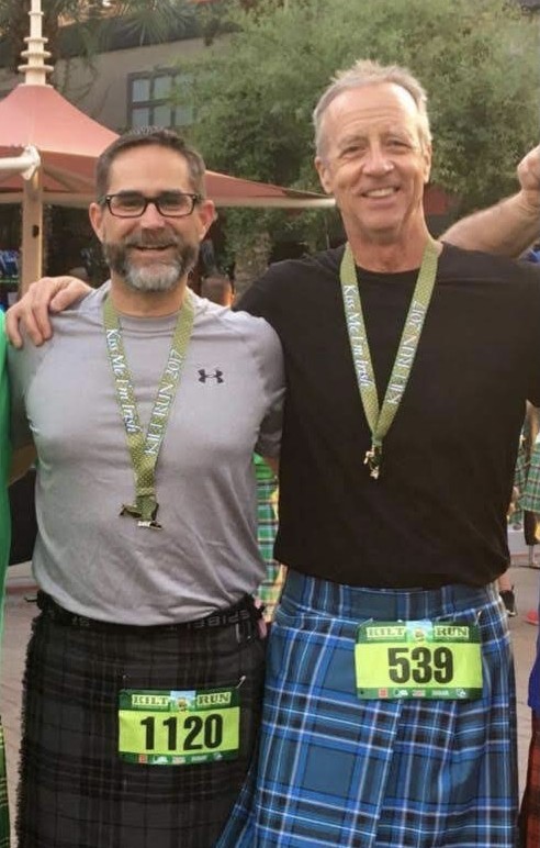 Two men stand outside, wearing kilts and exercise shirts, with race numbers pinned to their kilts. They're posing with their arms around each other