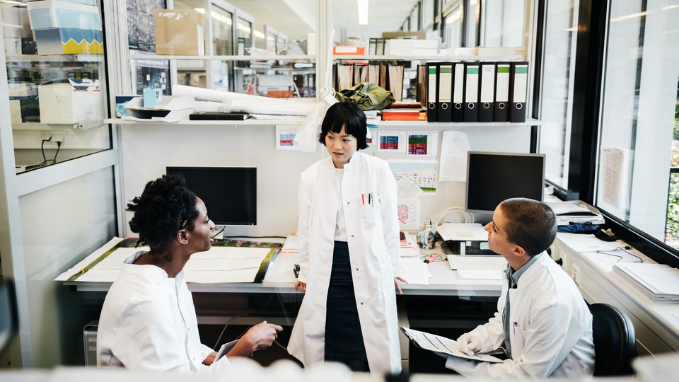 Three people in lab coats converse in a lab space with computers.