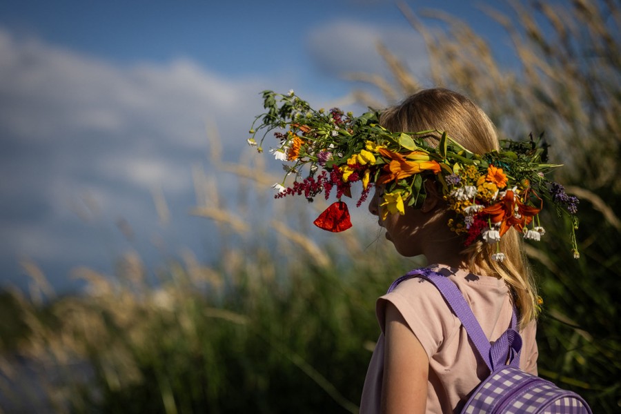A girl wearing a wreath on her head stands among reeds near a river.